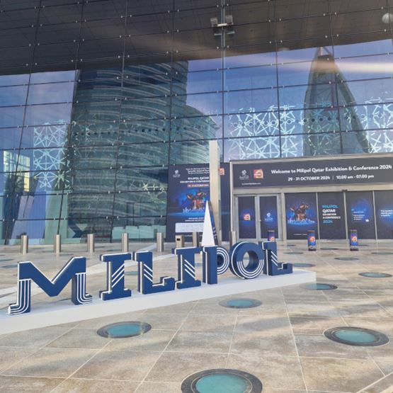 Exterior view of the Milipol Qatar 2024 exhibition center entrance with large Milipol sign in front and glass facade reflecting nearby skyscrapers
